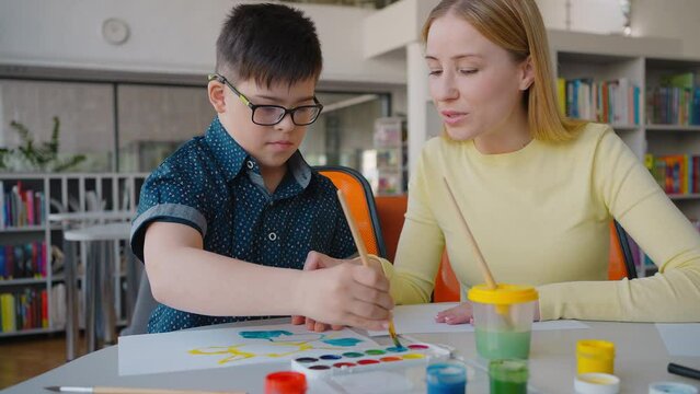 Happy elementary age boy with Down syndrome drawing watercolor picture during art class with teacher