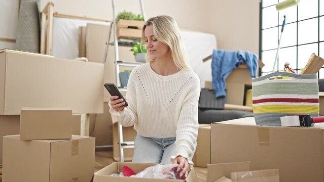 Young blonde woman unpacking cardboard box using smartphone at new home