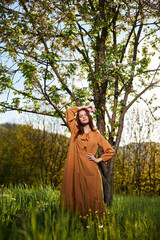 a sweet, attractive woman with long red hair stands in the countryside near a flowering tree in a long orange dress and looks at the camera holding her hand over her head
