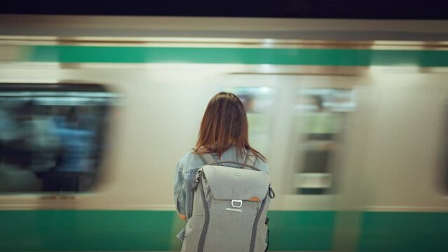 Rear view of young woman with backpack while waiting at platform of subway train station looking at passing tram, slow motion, waiting for a trip.