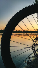 The silhouette of a mountain bike wheel on a river bank against the colourful backdrop of a sunset. An orange summer sunset over a river and a bicycle. Close-up.