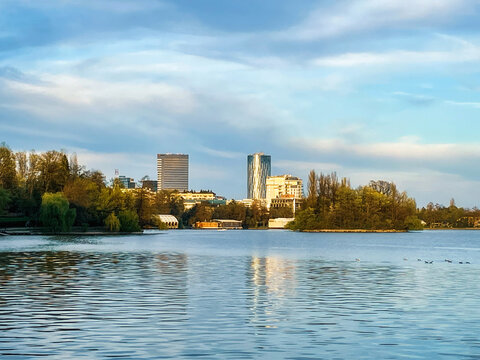 Scenic landscape view of Herastrau Park with trees reflected in the water on the edge of the lake at sunset and modern buildings in the background, in Bucharest, Romania.