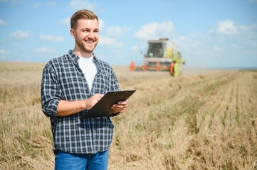 Handsome farmer with tablet standing in front of combine harvester during harvest in field.