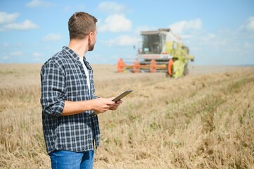 Handsome farmer with tablet standing in front of combine harvester during harvest in field.