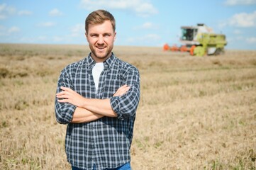 Happy farmer proudly standing in a field. Combine harvester driver going to crop rich wheat harvest. Agronomist wearing flannel shirt, looking at camera on a farmland