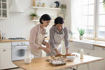 Positive adult daughter helping mature mom to bake, rolling raw dough on kitchen table, preparing homemade dessert, bakery meal, sharing domestic household chores