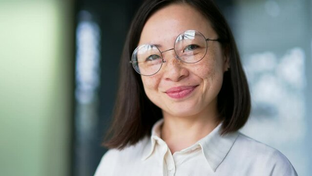Close up. Portrait of an adult Asian female employee standing in a modern office. Happy businesswoman in glasses is posing for a headshot. Successful manager, programmer, lawyer or woman entrepreneur