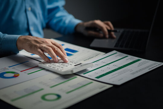 A businessman is using a white calculator to calculate financial numbers, he is sitting in his private office, the businessman examines the financial data from the corporate finance chart.