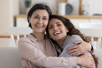 Cheerful positive mature 50s mom hugging adult daughter woman with head touch, relaxing on home sofa, looking at camera, smiling, laughing, enjoying closeness, family bonding, motherhood