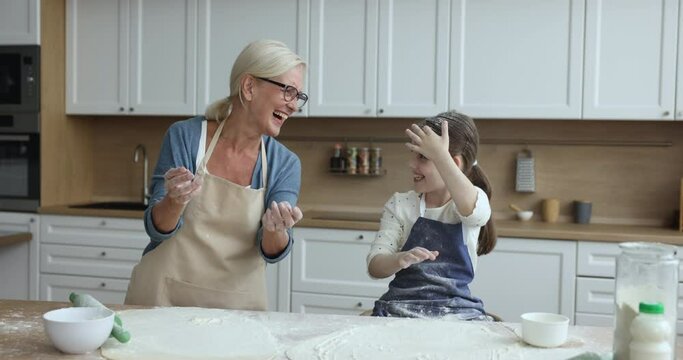 Happy mature grandmother and little granddaughter in aprons having fun, baking together, preparing dough clapping hands sprinkling white flour, laughing enjoy family time cooking holiday pie at home