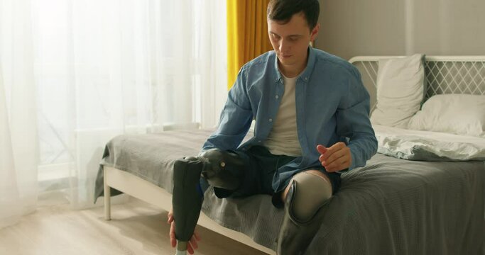 active ambitious disabled man putting on trainers, sneakers going to go in for sport Slow motion young man leg amputee wearing or changing prostheses at home sitting on bed in bright room, alone.