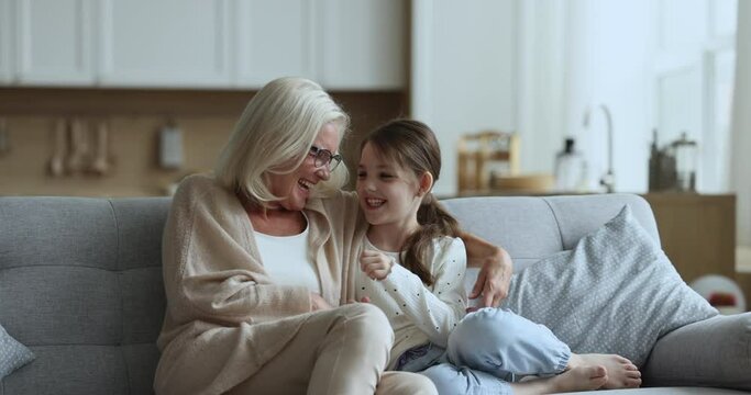 Happy middle-aged granny spend time weekend with adorable small granddaughter at living room, sit on cozy couch, tickle little girl enjoy leisure time together at home. Family ties, warm relationships