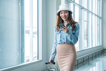 Asian female passenger walks holding a smartphone searching for travel information by herself dragging a suitcase while waiting for a connecting flight in the airport terminal.