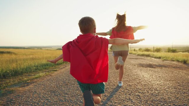 Carefree children play superheroes wear red cloaks run along rural road on nature with spreading flying hands.Teen girl sister kid boy brother have game together. Happy childhood, siblings friendship.