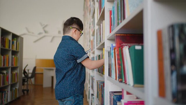 Child boy with Down syndrome selecting book from bookshelf in the library campus. Social integration