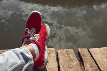Person in red shoes relaxing on a lake wooden deck. - obrazy, fototapety, plakaty
