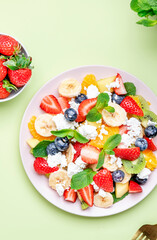 Summer fruit and berry salad with strawberries, blueberries, banana, soft cheese and mint leaves, green background, top view