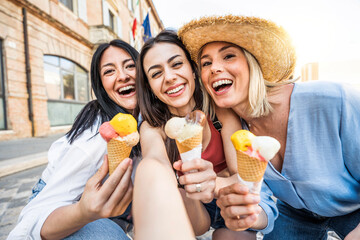 Fototapeta premium Three cheerful teenage women eating ice cream cones on city street - Happy female tourists enjoying summer vacation in Italy - Laughing girl friends taking selfie picture outside - Summertime holidays