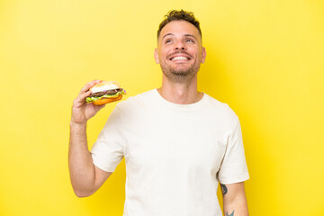 Young caucasian man holding a burger isolated on yellow background thinking an idea while looking up