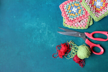Red scissors, cotton yarn balls, crochet hook and granny squares on a blue desk. Messy working place. Crochet geometric shape. 