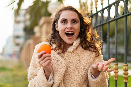 Young caucasian woman holding an orange at outdoors with shocked facial expression