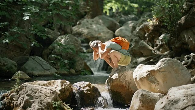 slow motion man hiker walks through river by boulders and drinks fresh cold water from stream during hike in jungle. hike in wild forest at hot summer day, outdoor activity and travel concept