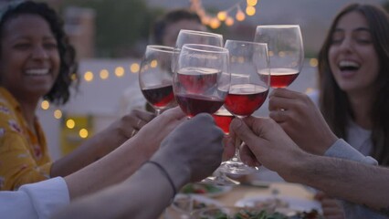 Gathering of cheerful young friends toasting with red wine and celebrating party on terrace of house. People together drinking alcoholic beverages outdoor. Boys and girls enjoying free time on weekend