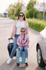 Adorable little girl sits on her kid suitcase and waiting for the trip with her mom. Two beautiful girls prepare to journey. A car is parked on the side