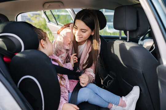 Attractive Young Mother Put Her Daughter In A Car Seat And Fastens Her Seat Belts. Woman Care About Kid. Protection During The Trip In The Car.