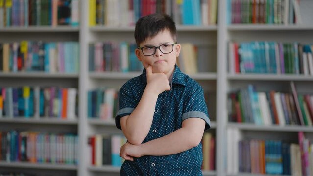 Pensive schoolboy with Down syndrome dreamily looking aside, standing over bookshelves background
