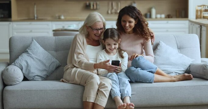 Three happy carefree generations of women relax at home on cozy sofa in living room with cell phone, watch funny online content or video, use modern application, enjoy pleasant family leisure at home