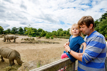 Cute adorable toddler girl and father watching wild rhinos in zoo. Happy baby child, daughter and dad, family having fun together with animals safari park on warm summer day. Ireland