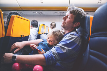 Young father holding his baby toddler daughter during flight on airplane going on vacations. Tired...