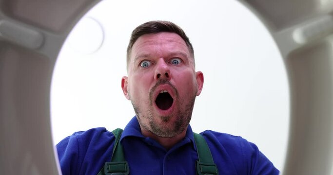Shocked male plumber opens toilet lid putting hands on head. Man prepares to clean clogged sewer pipe. Expressive person showing surprise