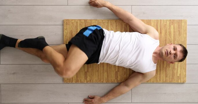 Man does relaxing exercise lying on reed matting put on laminate floor in light premise. Athletic man enjoys effective training at home upper view
