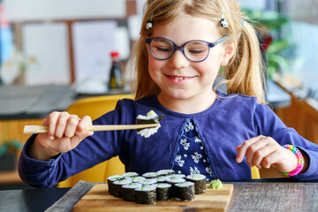 Happy preschool girl eating sushi rolls in a cafe or restaurant using chopsticks. Healthy food for children and youth. Preteen child tasting japanese food. Little child with glasses enjoying meal.