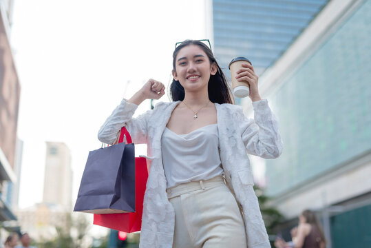A cheerful asian female office employee feeling optimistic while walking around the city district. Holding a cup of coffee and some shopping bags.