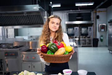 Portrait of a female student chef holding a basket of fresh vegetables in the kitchen.