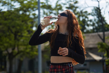 Thirsty young adult woman with long red hair drinking water from a reusable glass bottle outdoor. Portrait of a white female person in sunglasses hydrating herself with a sip of fresh water