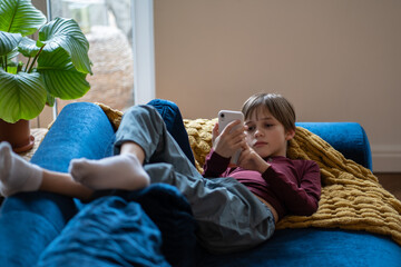 Depressed sad little kid schoolboy lying on sofa looking on mobile phone screen feeling upset,...