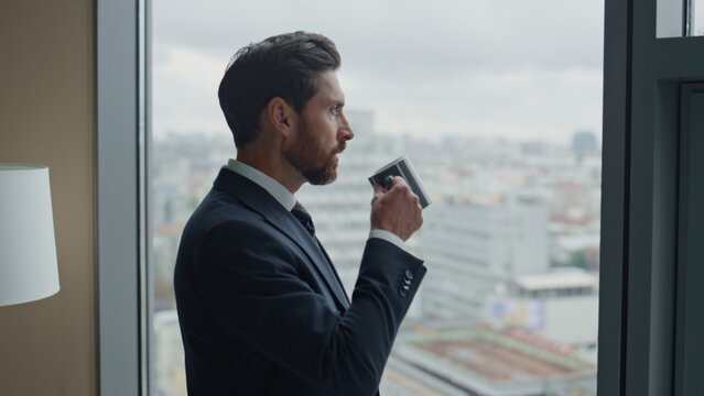 Pensive boss drinking coffee at window office watching cityscape alone close up.