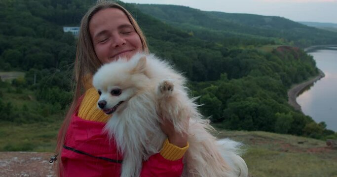 Cute happy adult woman hugging spitz dog sitting on peak of mountains, river in the background, Slow motion backpacker embracing pet, love friendship concept girl takes care of Spitz