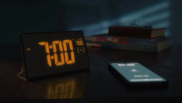 Digital alarm clock with orange clockface and the smartphone waking up at 7 AM. The numbers on the clock screen changes from 6:59 no 7:00 AM. And the alarm goes off on the phone. Close up view.