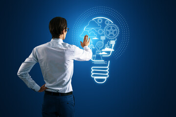 Back view of businessman hand using abstract digital lamp hologram on blue background. Start up, entrepreneurship and creative ideas concept.