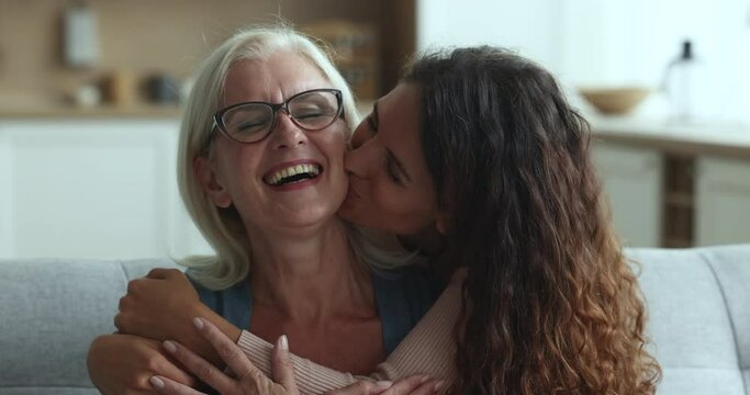 Loving grown up daughter cuddling and kissing on cheek her happy senior mum miss after long separation express love, enjoy tender moment sit on sofa during her visit, close up. Motherhood, family ties