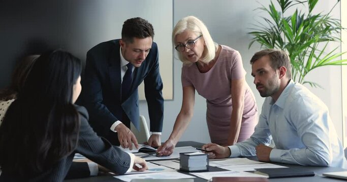 Serious different aged business leaders discussing paper sales report, marketing strategy on corporate meeting, standing at table, pointing at documents, speaking. Senior coach training group