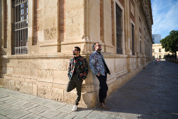 Young fashionably dressed gay male couple are leaning against the corner of a historic building, each looking to one side. They are on a honeymoon vacation. The couple are married. Gay rights