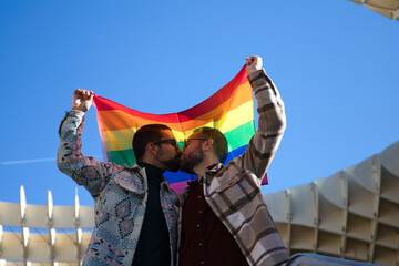 A young couple of gay men holding the gay pride flag on their backs. The couple kiss and are happy. Gay rights concept