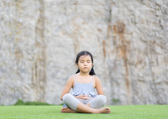 Asian child meditate zen or kid girl close eye mind meditating with breath and sitting on meadow green grass for peace yoga to hand overlap meditation at outdoor temple park and stone wall background