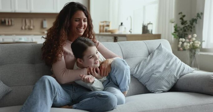 Cheerful young mother tickles daughter spending time together at home sit on cozy couch, have fun on weekend, playing active games, laugh, mom cuddling child, enjoy happy motherhood and entertainment